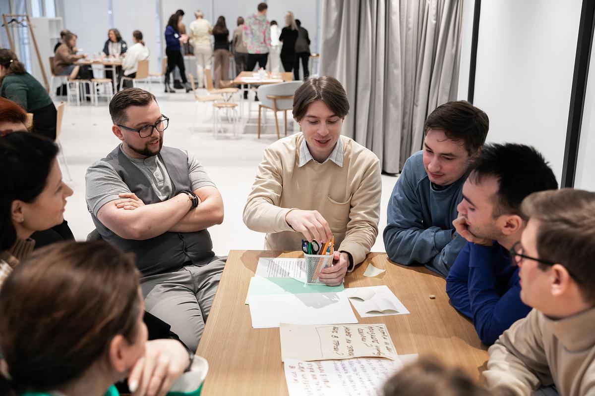 Титульное изображение для страницы события: группа людей сидит за столом и что-то обсуждает, на столе разложены бумаги с записями