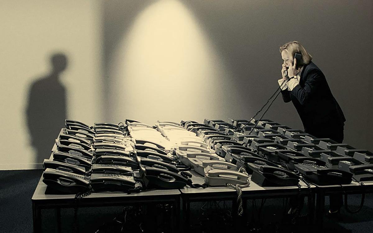 Титульное изображение для страницы события: кадр из фильма «Белый квадрат», женщина в черном костюме стоит перед столом с огромным количеством телефонов и разговаривает по двум из них