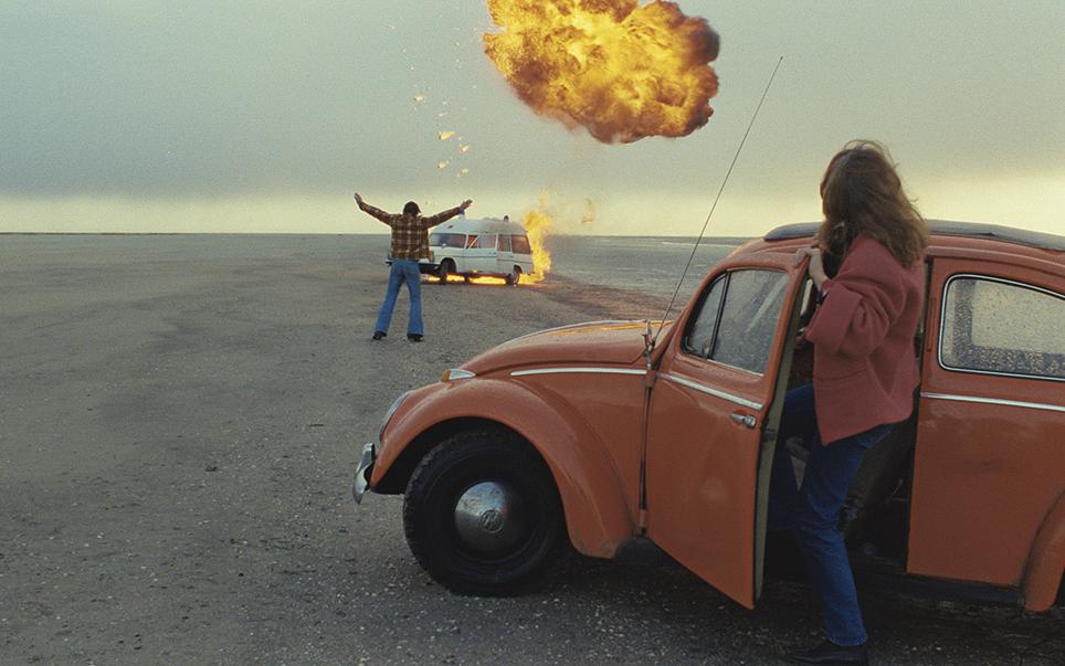 Титульное изображение для страницы события: кадр из фильма «Американский друг», на переднем плане женщина выходит из машины и смотрит вдаль на мужчину, вздымающего руки на фоне горящей машины