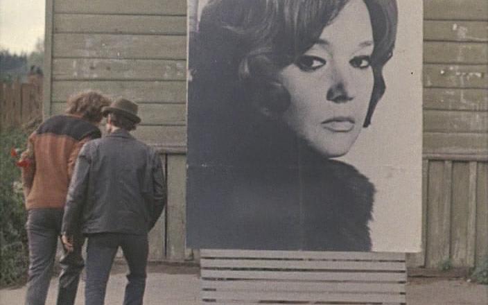 Титульное изображение для страницы события: кадр из фильма «Познавая белый свет», два человека идут по улице мимо большого портрета женщины на деревянной стене