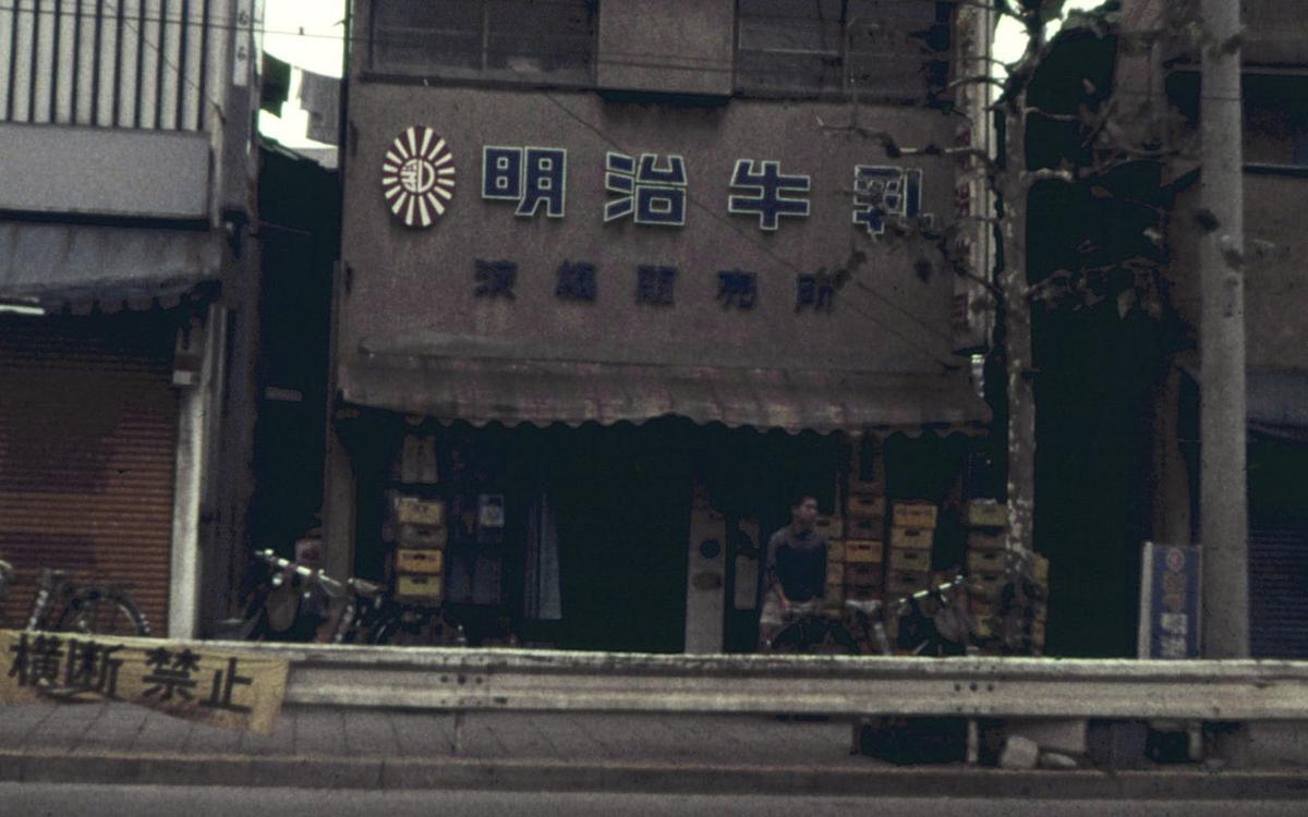 Титульное изображение для страницы события: кадр из фильма «Известен также как серийный убийца», человек сидит у входа в задние с японскими надписями на фасаде