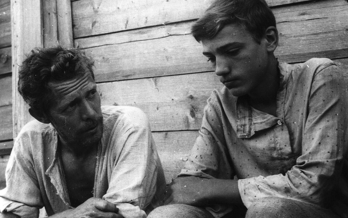 Титульное изображение для страницы события: кадр из фильма «Начало неведомого века», два мужчины сидят на фоне деревянной стены, один смотрит вперед, второй - на первого
