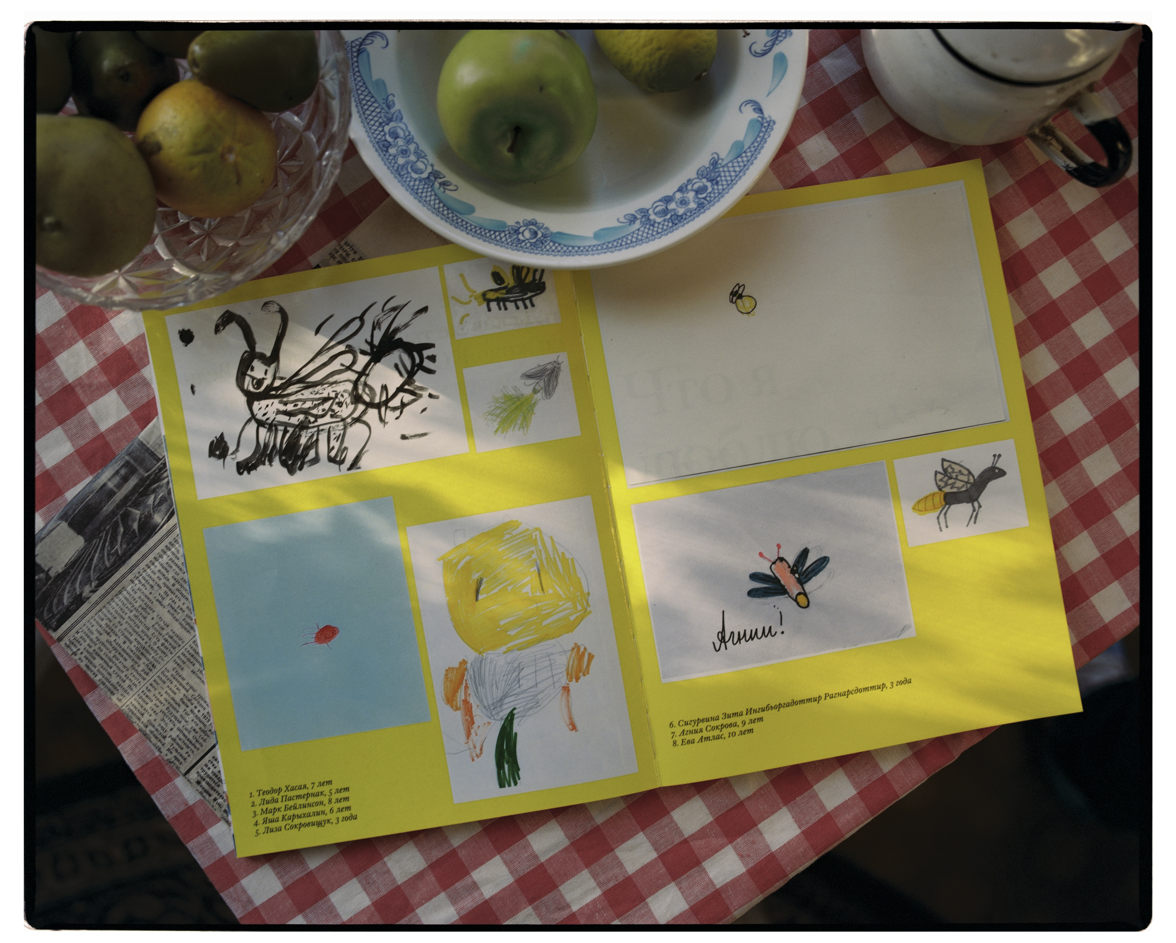 Разворот книги с детскими рисунками, лежит на столе со скатертью в красно-белую клеточку