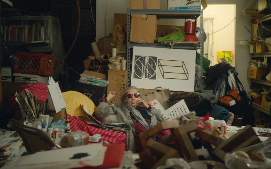 Человек в шубе и солнцезащитных очках лежит на груде мусора посреди комнаты
