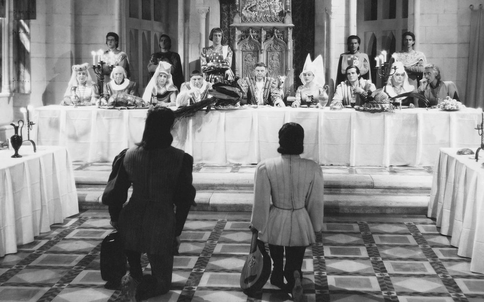 Два менестреля склонили колено перед столом, за которым сидят представители аристократии