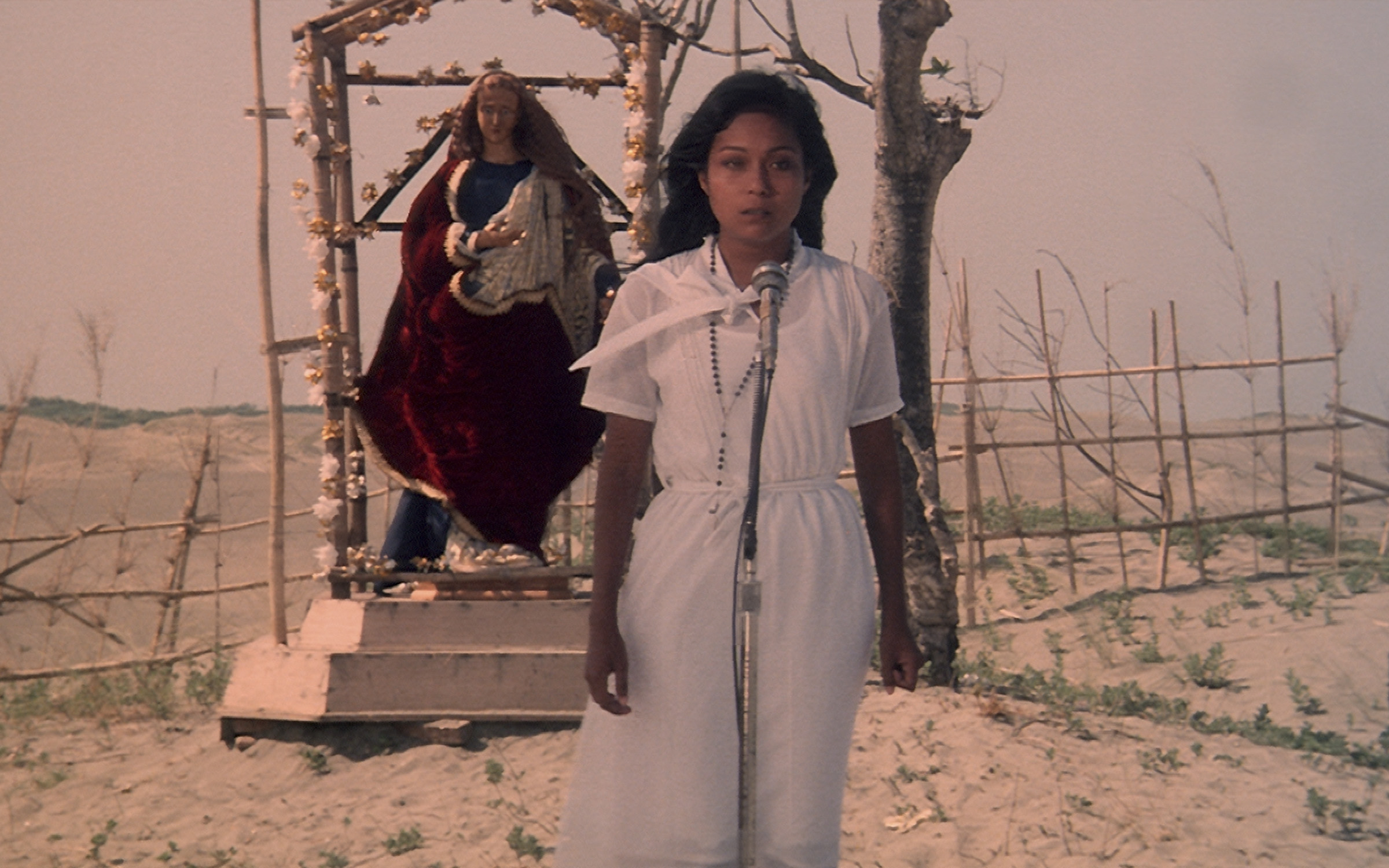 Женщина в белом стоит на фоне статуи Девы Марии в пустыне