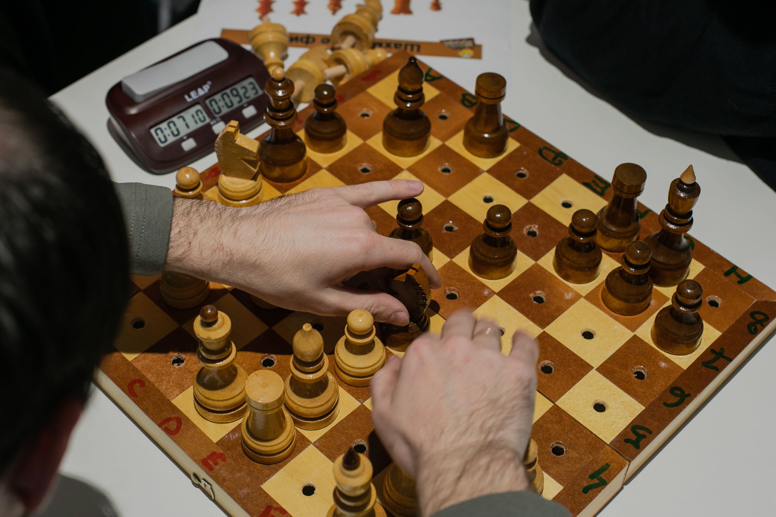 Цветная фотография: на белом столе раскрыта шахматная доска, рядом с которой лежит таймер.  Над доской нависли руки мужчины, который переставляет фигуры. Доска усеяна отверстиями, которые нужны для фиксации фигур