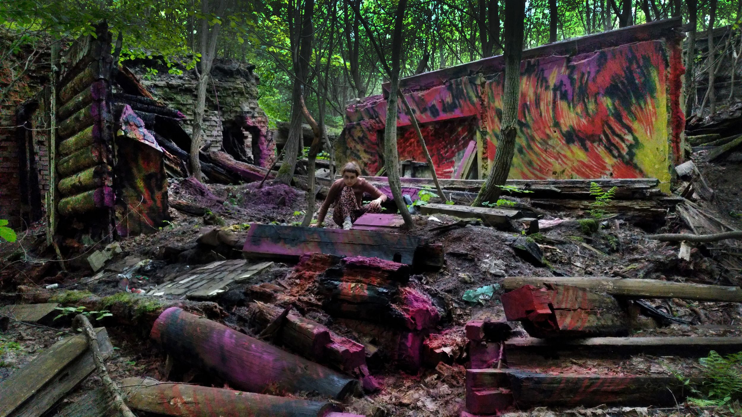 Фотография деревянных развалин в лесу, стены украшены яркими желто-красными пламевидными граффити, в центре на корточках сидит девушка